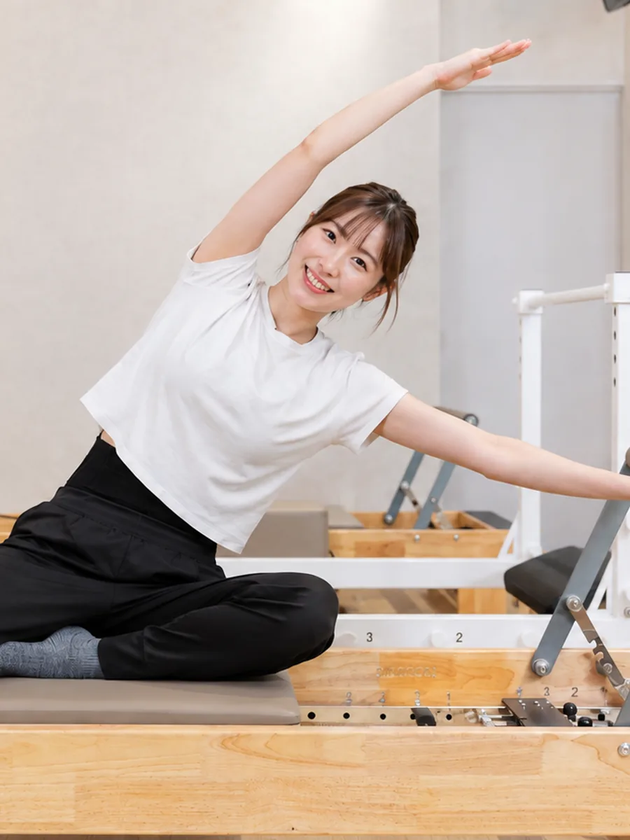 明るいスタジオでリフォーマーに座り体側を伸ばす女性